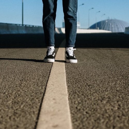 A lone individual stands on an empty divided road under a clear blue sky, emphasizing isolation.