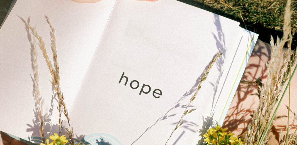pexels-photo-5063828-5063828 Hand holding an open book displaying 'hope' among yellow flowers in a field.